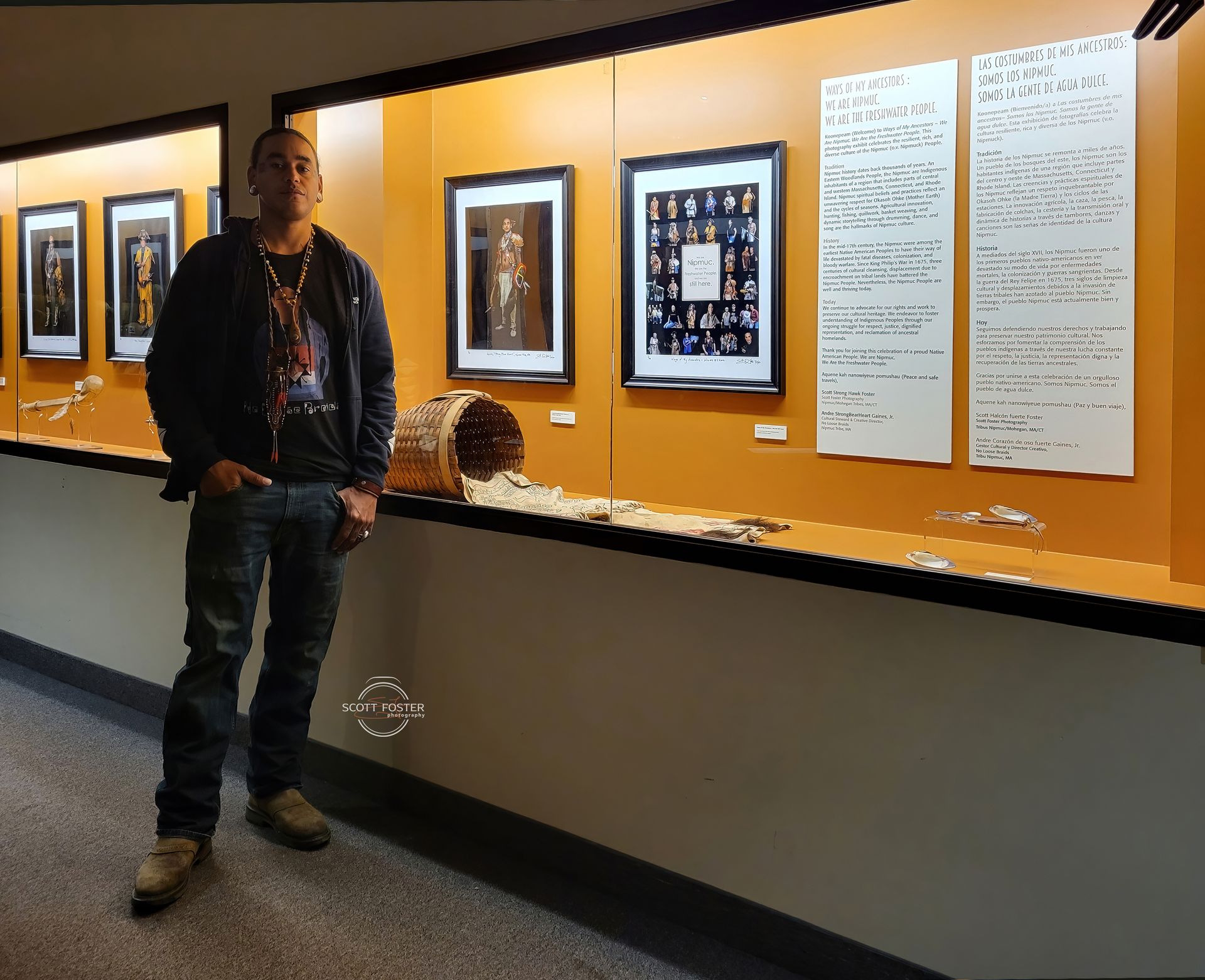 Person standing in front of a display case with framed art and artifacts, indoors. Orange wall, neutral tones.