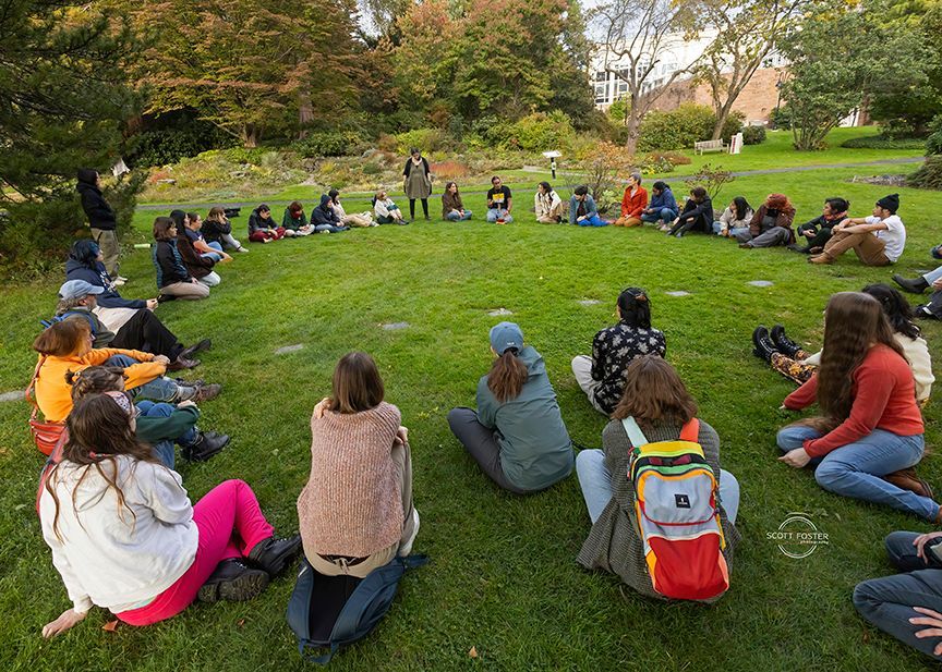 A group of people sit in a circle on grass, listening to a speaker in a park.