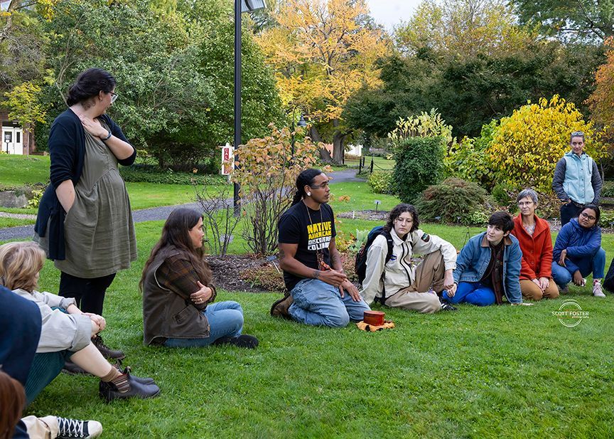 Group of people seated outdoors on grass, listening to a person kneeling with a small drum. Autumn trees in the background.