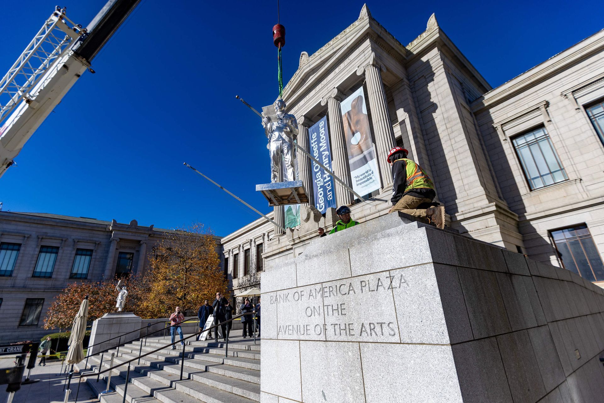 A crane lifts a statue above the Denver Art Museum, workers observe. Sunny day, blue sky.