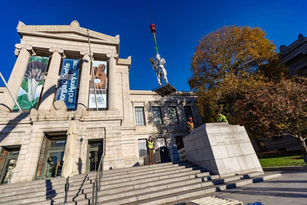 Construction workers lift a large statue with a crane, in front of a stone building with flags and a monument.