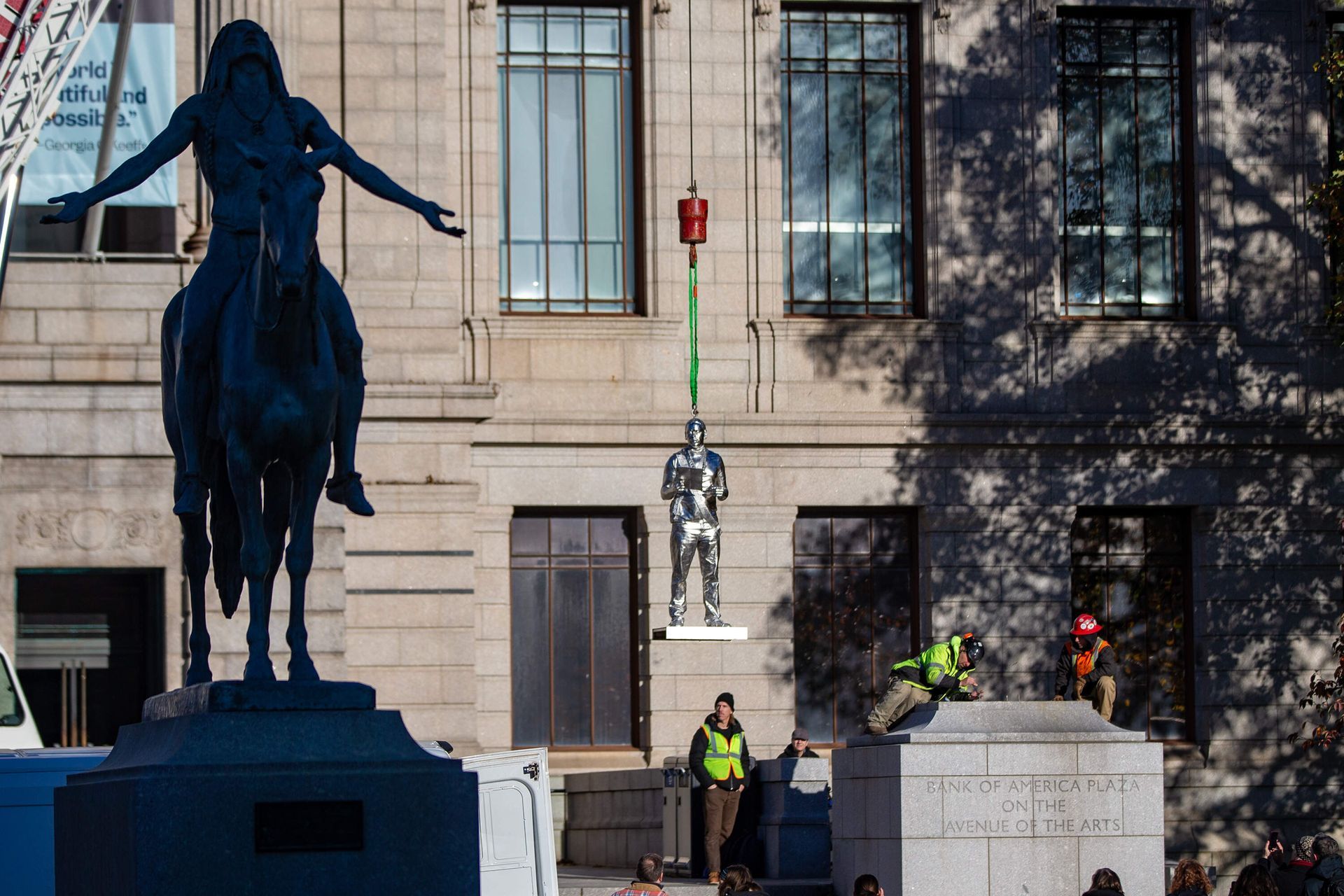 Statue of a man on horseback, workers removing a statue from building, crane lifts a sculpture. Sunny day.