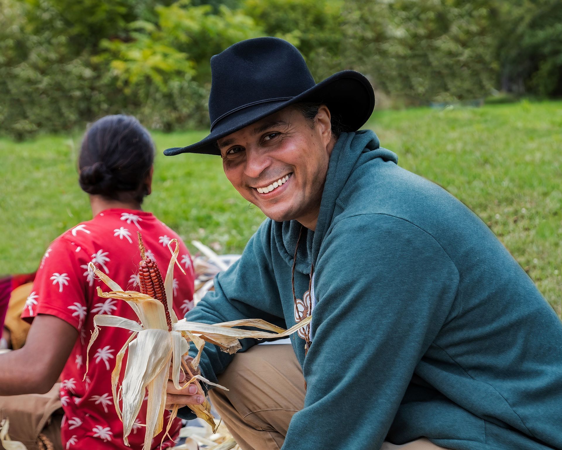 Man in cowboy hat smiles, holding corn cob, outdoors. Green sweatshirt, another person in red shirt.
