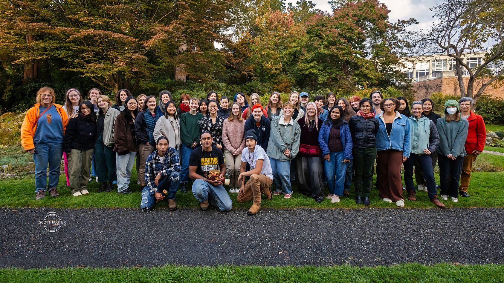 Group of people pose on grass. Autumn trees in the background.