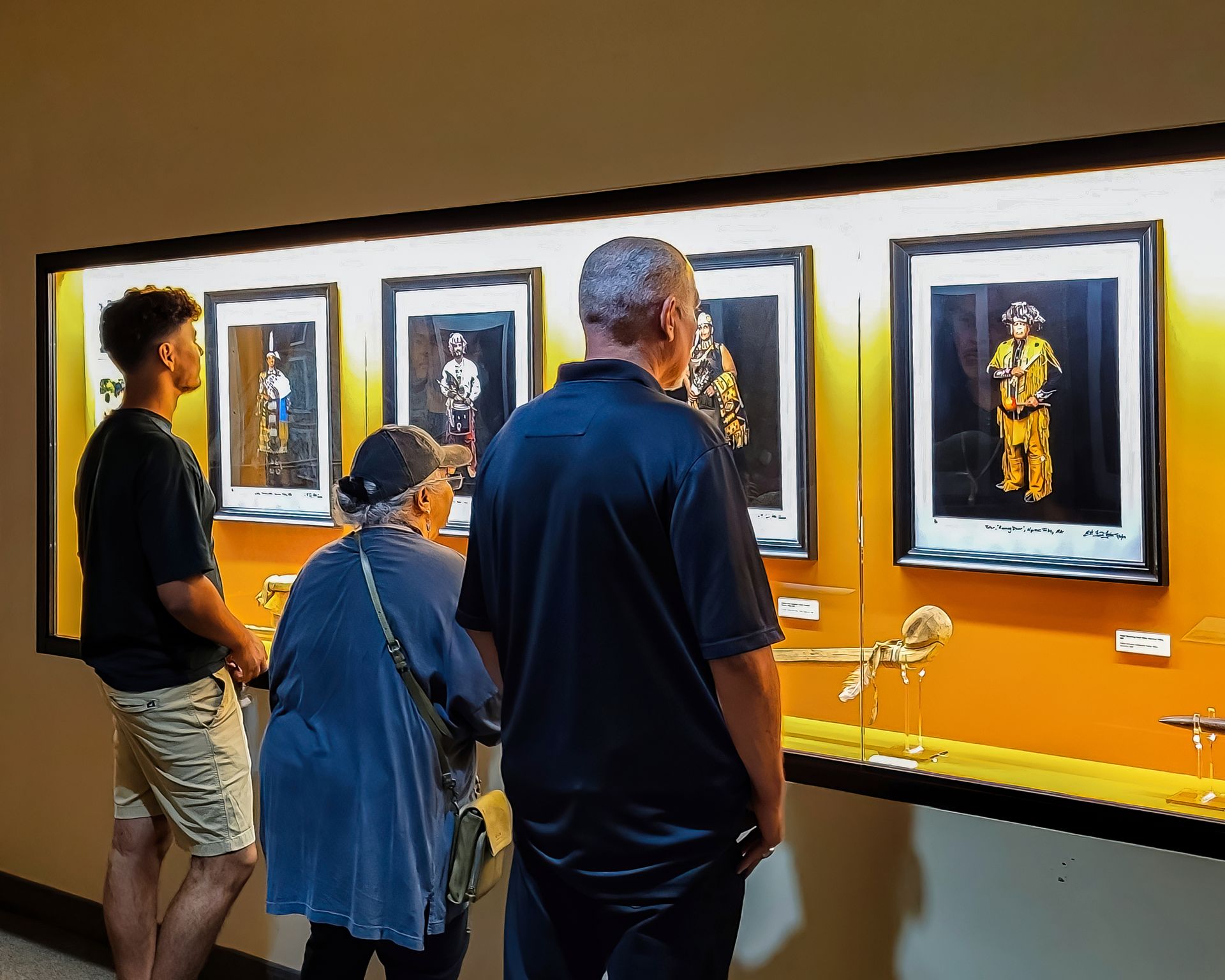 People looking at framed artwork in a museum. Yellow wall and display case with Native American images.