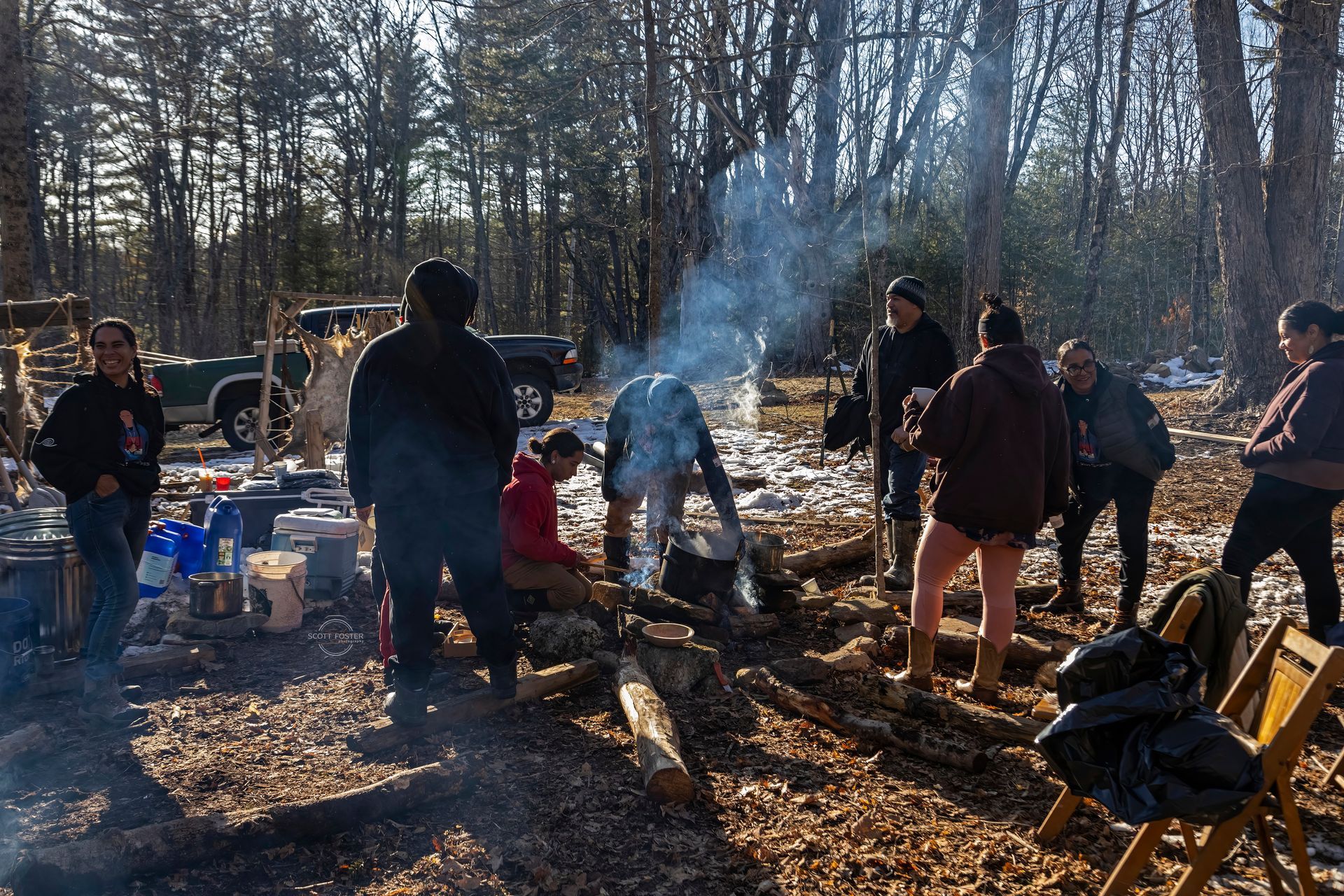 Group of people around a fire outdoors, preparing food. Smoke rises, trees in the background, a truck parked nearby.
