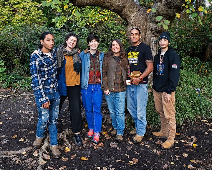 Group of six people smiling in front of a tree with fall foliage. They wear casual clothing and stand on fallen leaves.