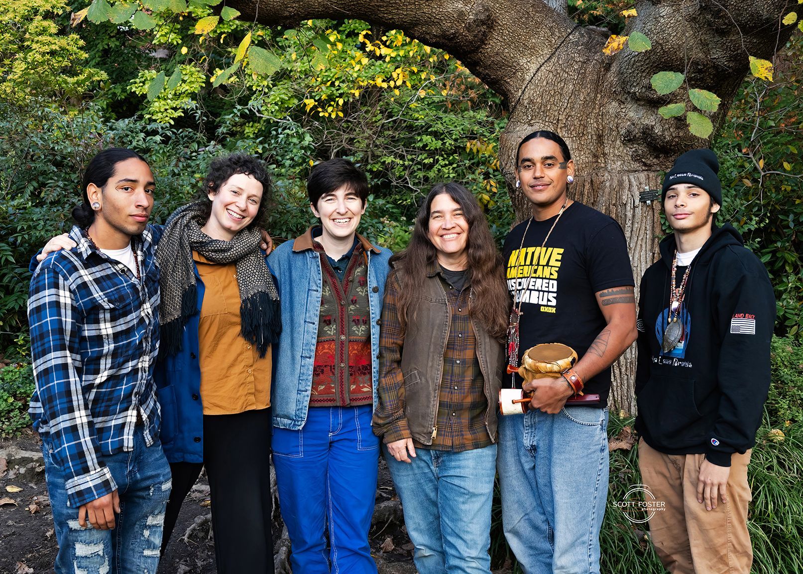 Group of six people smiling, posing near a tree with fall foliage. They wear casual clothing, outdoors.