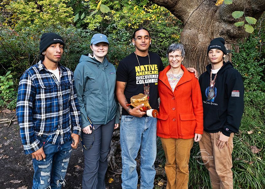 Five people pose outdoors by a tree. One holds an object, others wear hats and jackets.