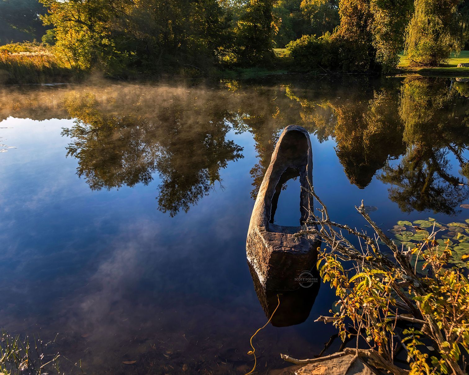 Stone sculpture in calm water, trees reflected. Haze above the water on a sunny day.