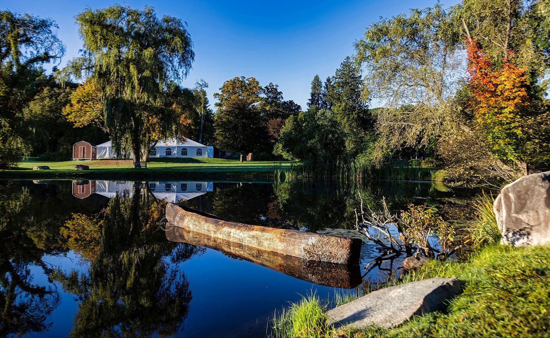 A still lake reflects trees and a white house, with an old wooden boat in the foreground.