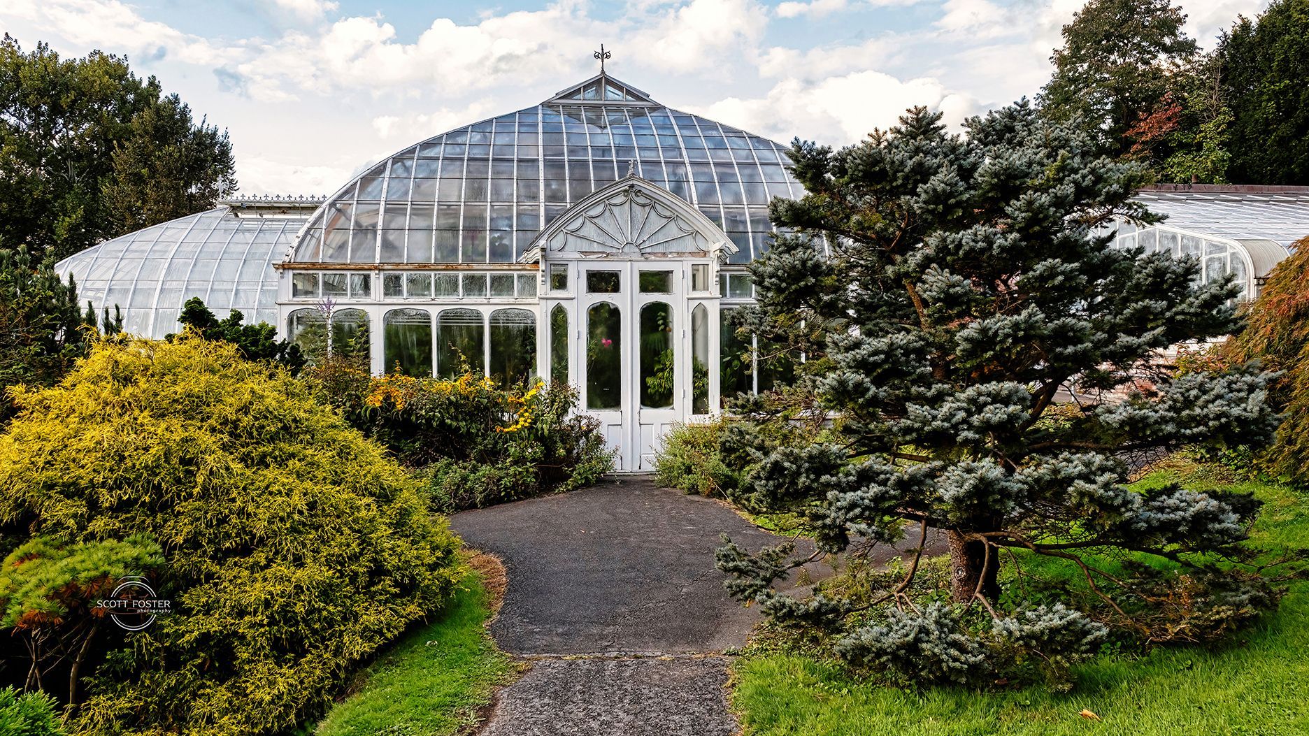 Glass conservatory with white trim and doorway, surrounded by green shrubs and trees, pathway leading to entrance.