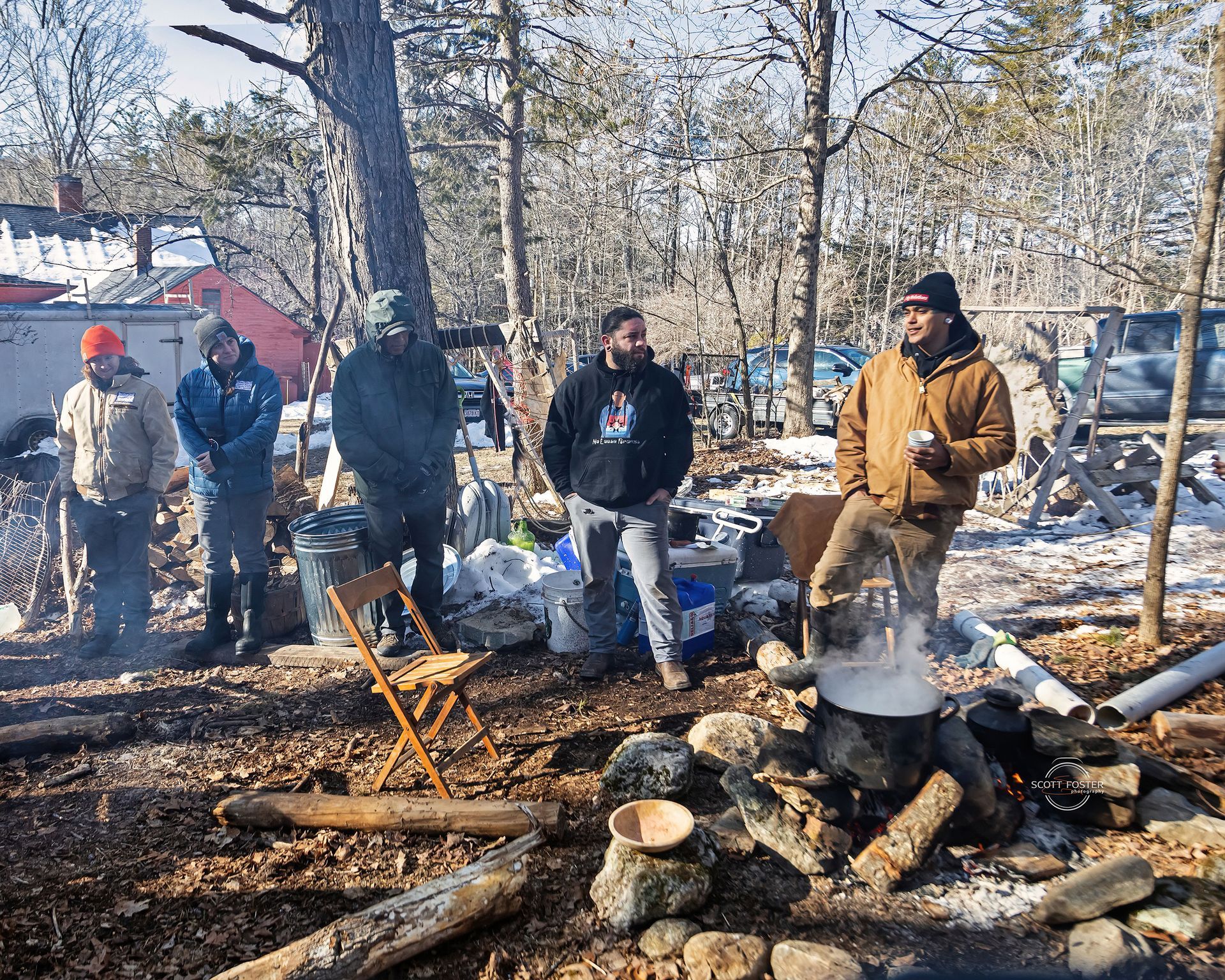 Men standing around a fire outdoors, near a small boiling pot. Winter setting, with trees and snow.