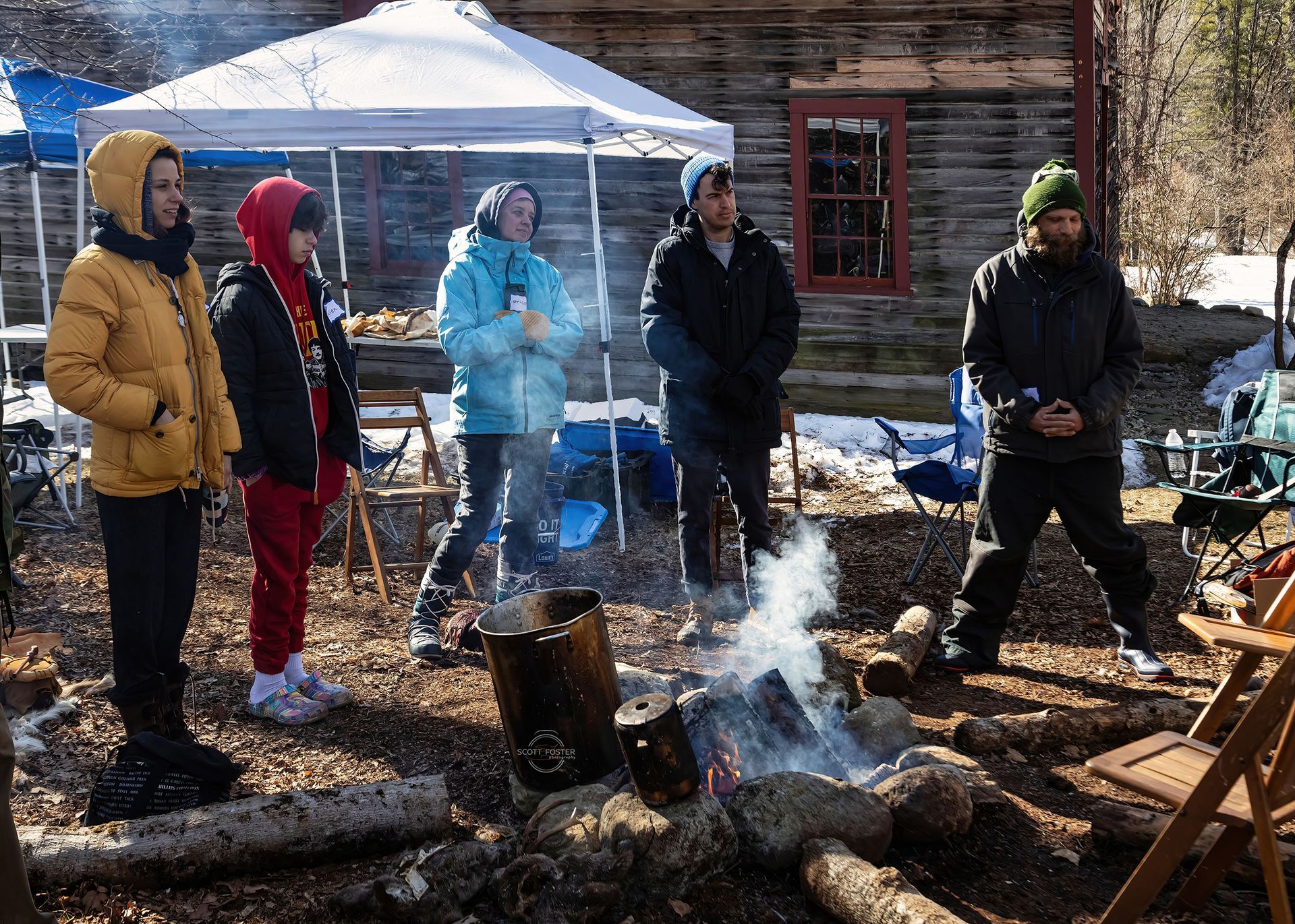 People gather around a campfire outside a wooden building. Snow on the ground.