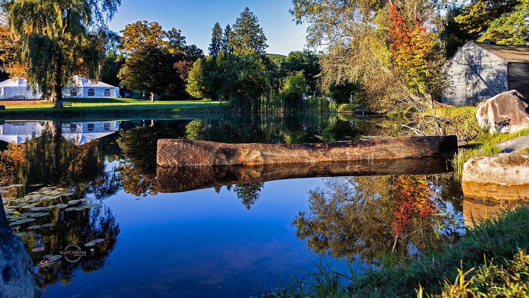 Calm pond with reflections of trees and buildings; a weathered stone structure floats in the center.