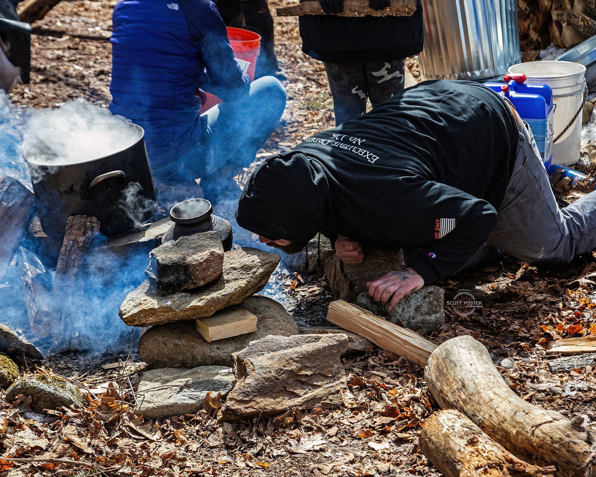 Person smelling a stone stack near a fire with a pot; outdoor setting with vapor.
