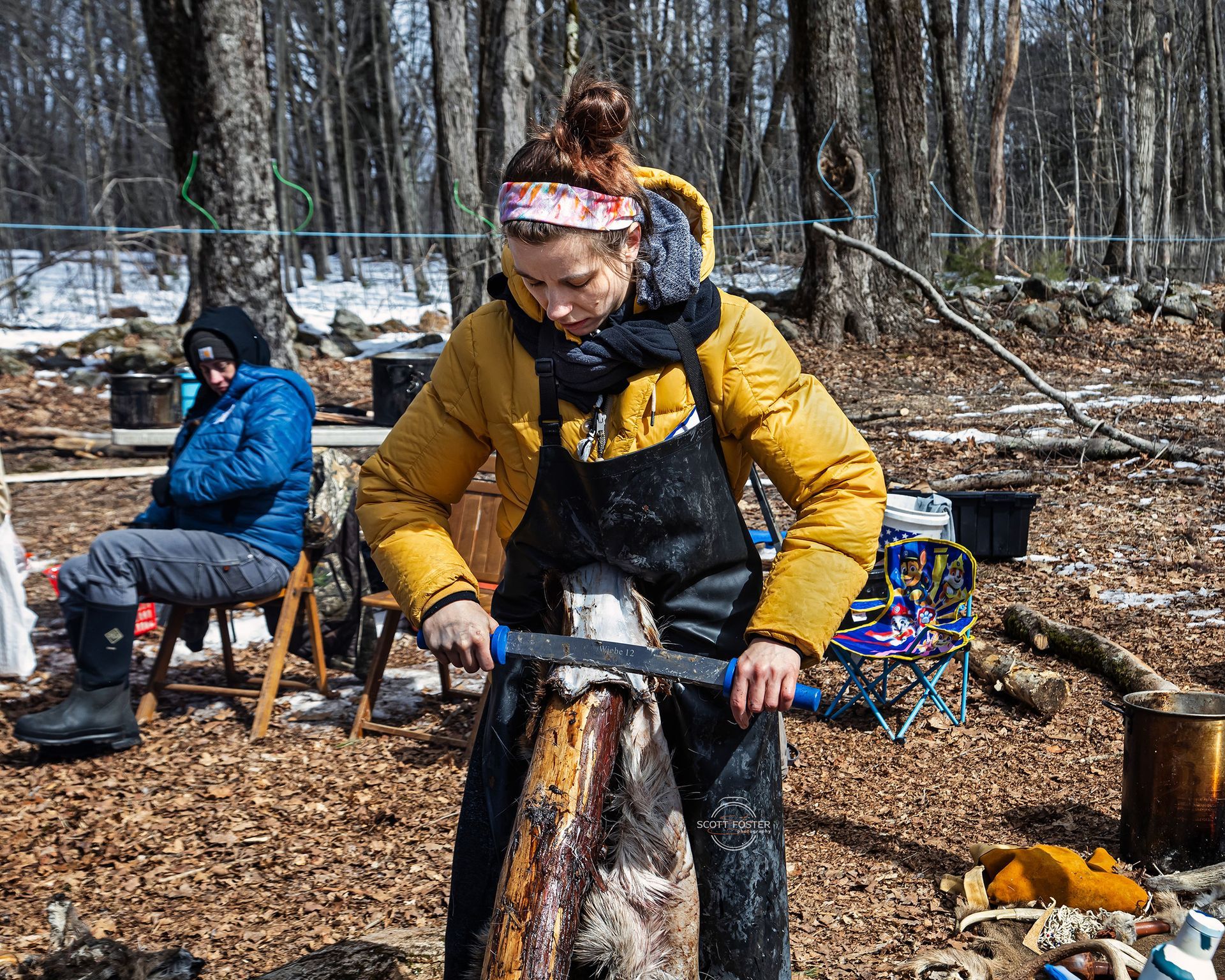 Woman in yellow jacket uses a knife to work on a large piece of wood outdoors. Another person sits in the background.