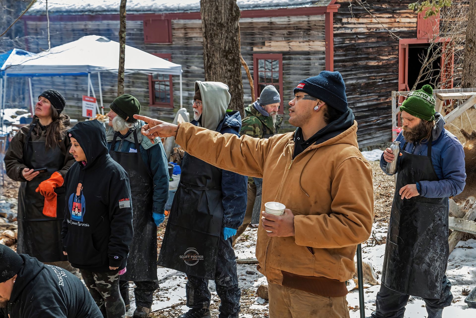 A group of people outdoors, listening to a person pointing. Snow on the ground, wood structures in the background.