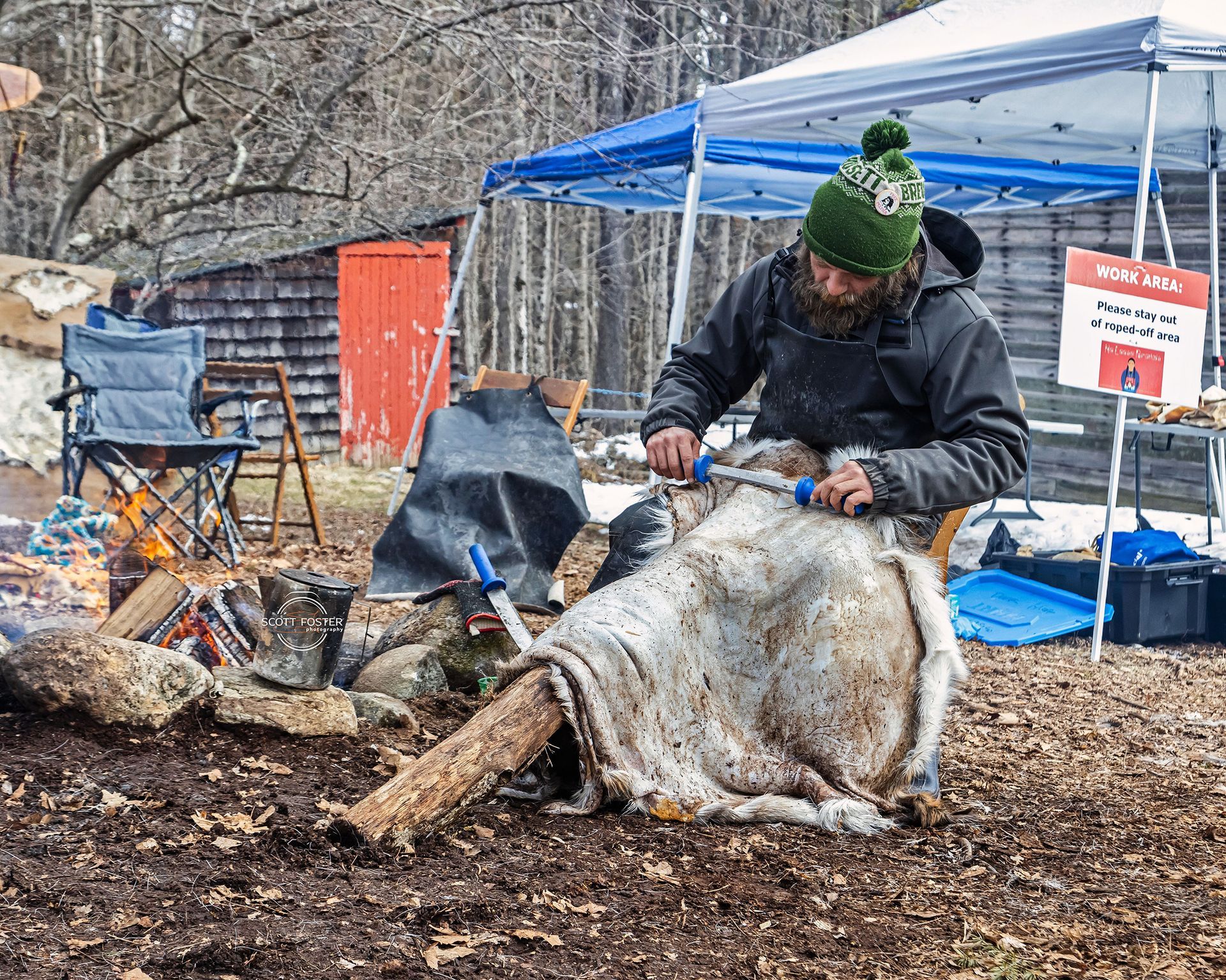 Man outdoors using tools to work on animal hide, near a fire and tents.