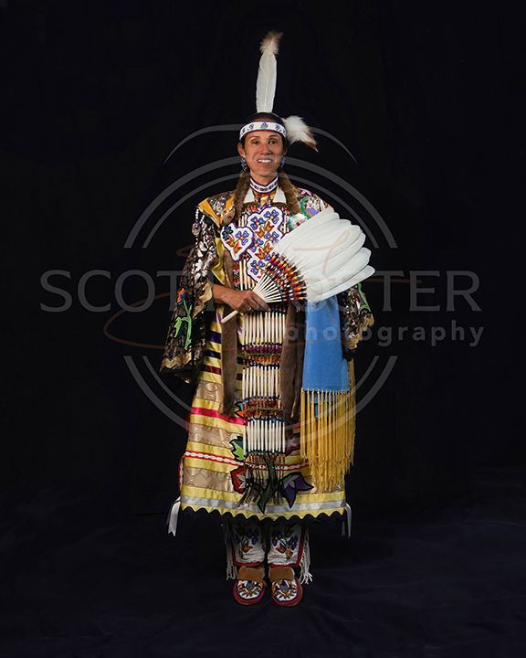 Woman in Native American regalia, holding a fan, smiling. Black background.