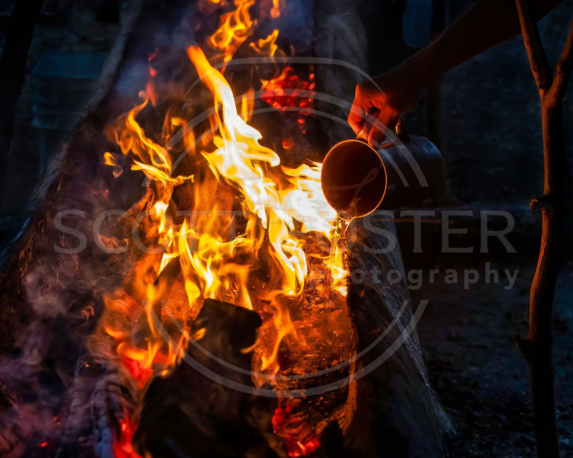 Fire being doused with water from a metal container, flames and wood visible, dark setting.