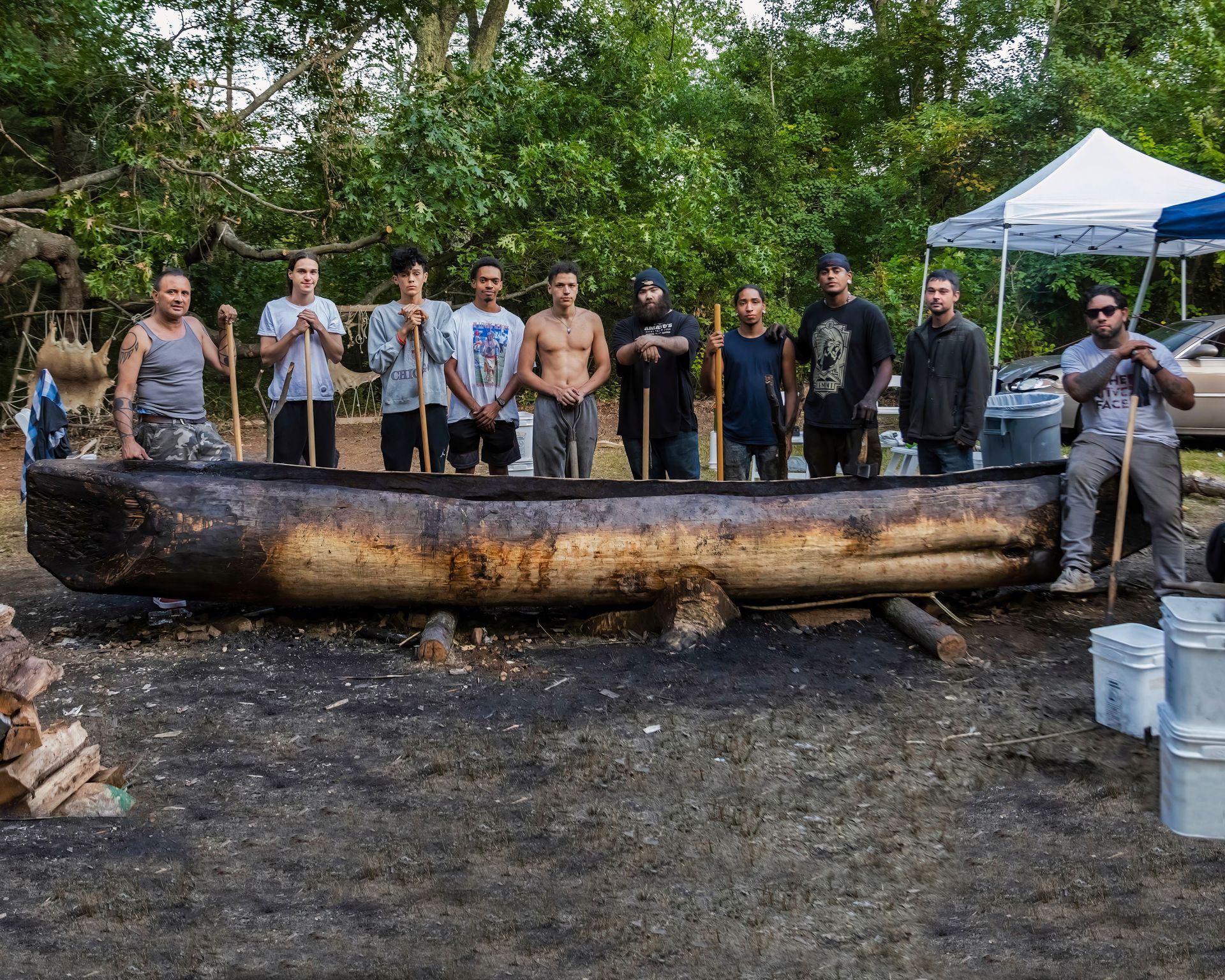 Men standing behind a large, dark canoe, holding tools. Outdoor setting with a tent and trees.