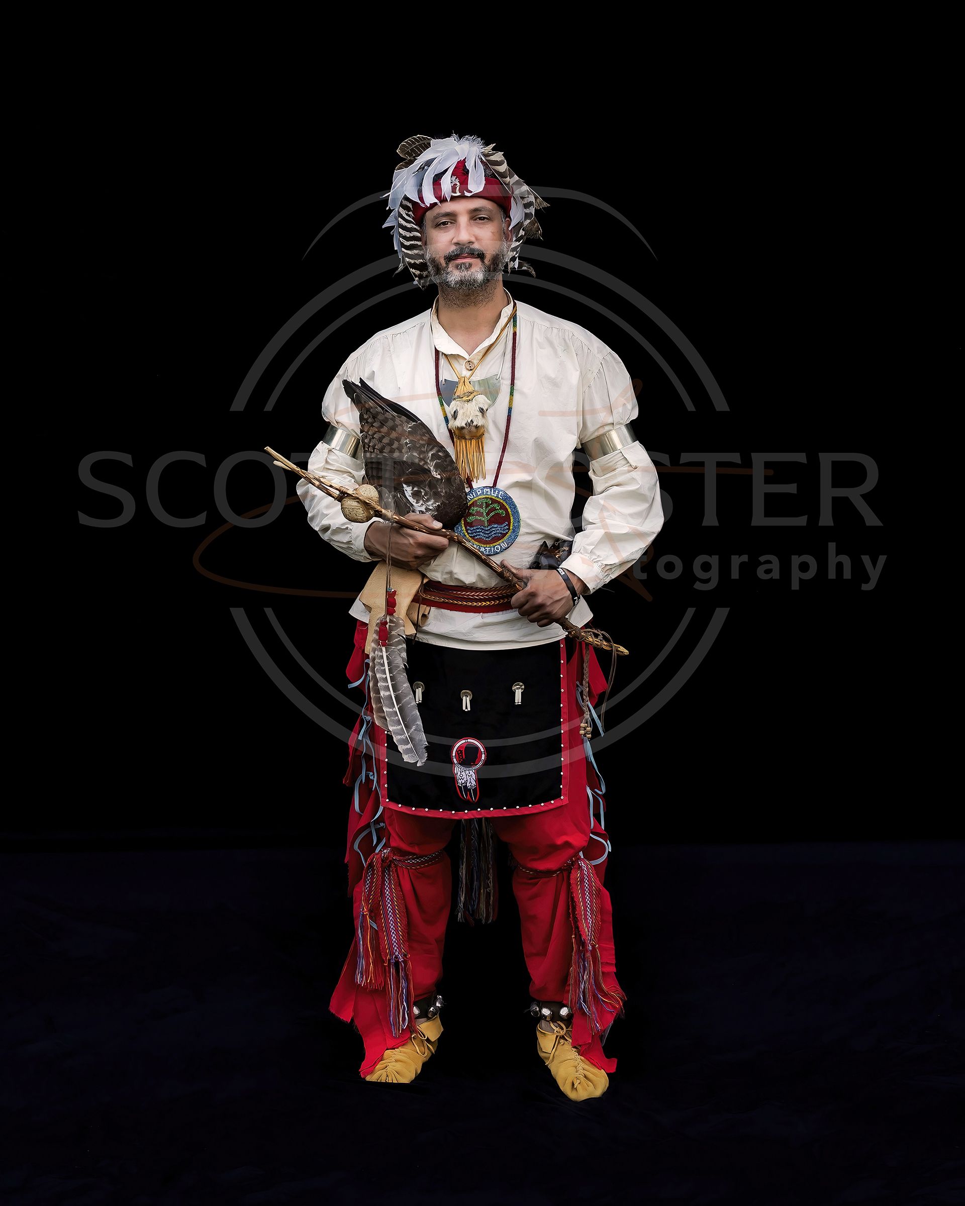 Man in traditional Indigenous regalia, holding a staff with an eagle. Black background.