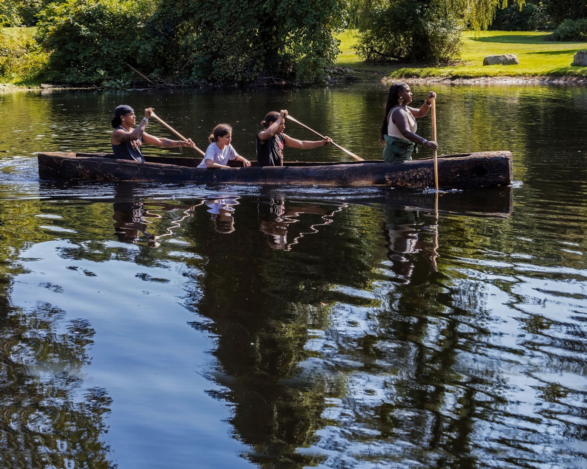 Four people paddling a wooden canoe on a calm lake. Trees and green vegetation surround the water.