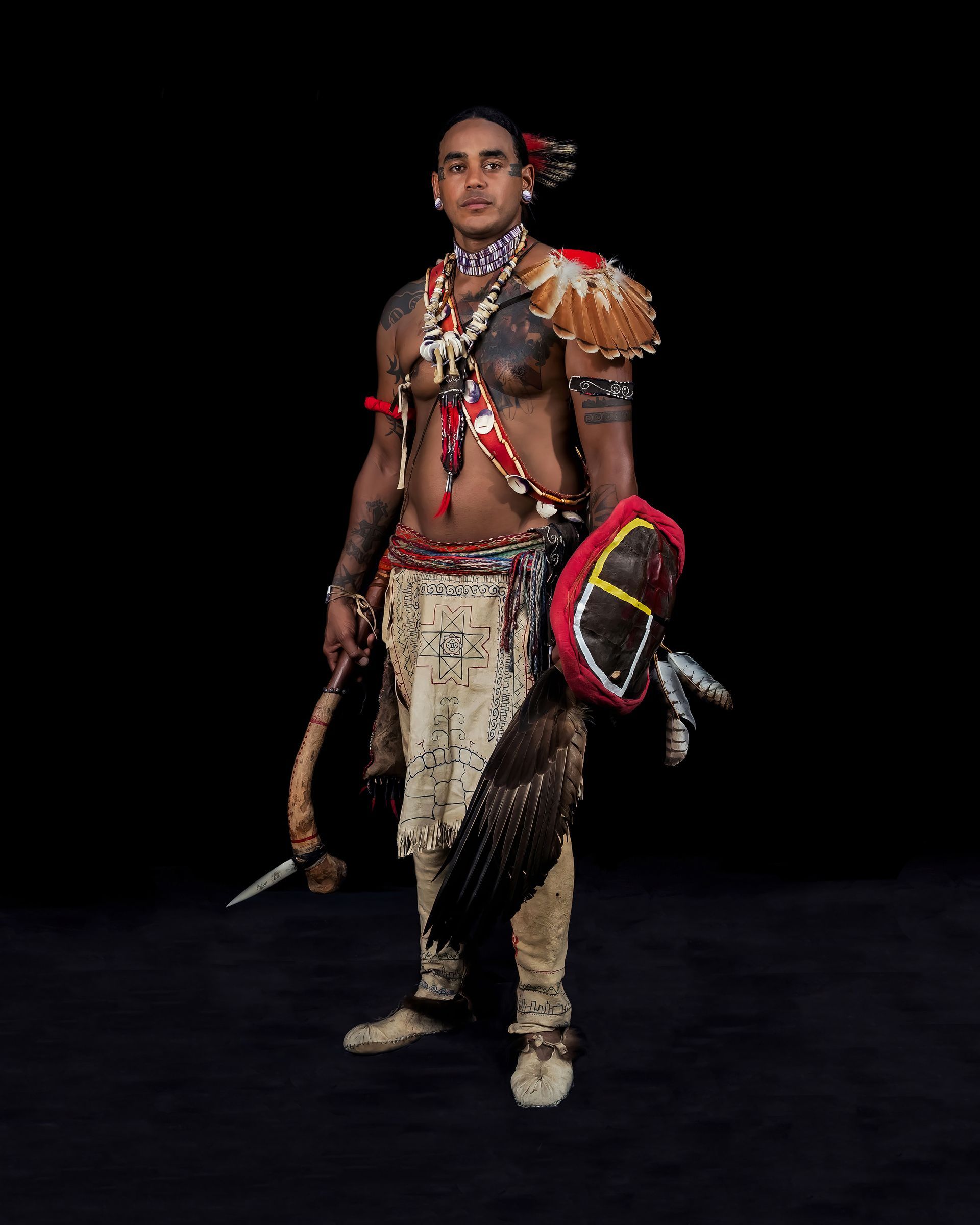 Man in Native American attire, holding a weapon and shield, against a black background.