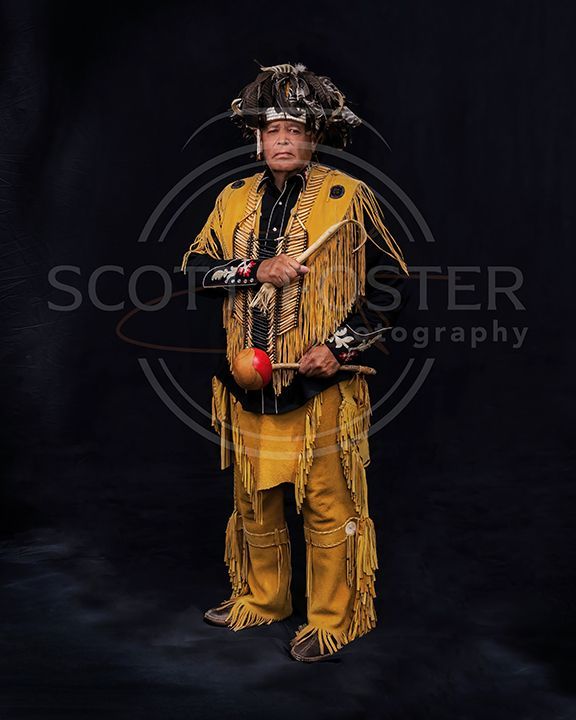 Man in yellow fringed regalia, holds drum with mallets, wearing headdress, dark background.