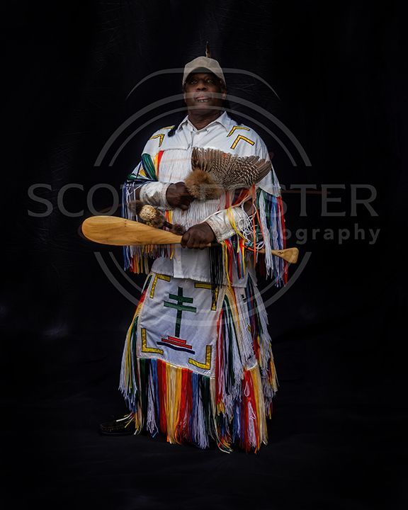 Man in ceremonial garb, holding a rattle and wooden paddle, standing against a dark background.