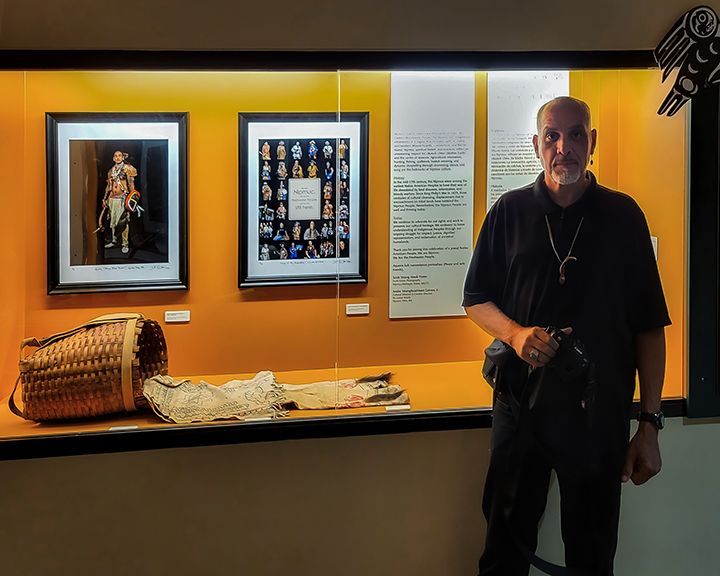 Man in a black shirt stands in a museum exhibit with framed art, artifacts, and a display case.