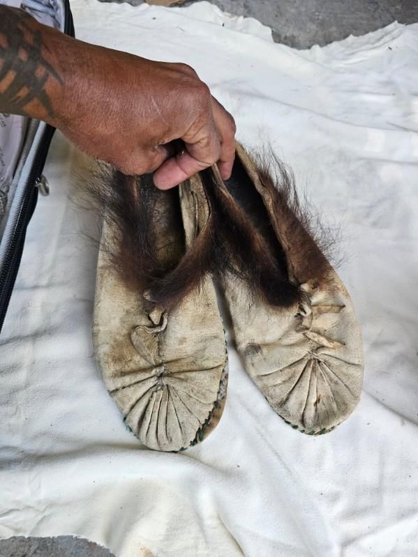 A hand holding two worn, fur-trimmed leather moccasins on a white cloth.