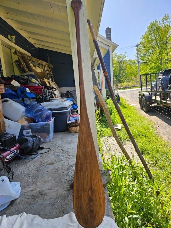 A large wooden paddle leaning against a porch, next to other wooden poles.