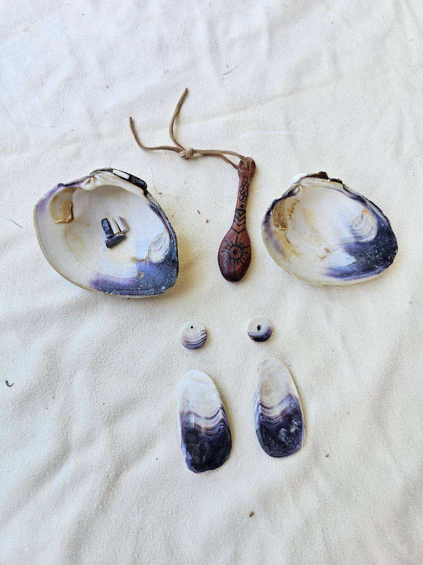 Seashells, a small wooden artifact, and beads on a sandy surface. Some shells have purple edges.