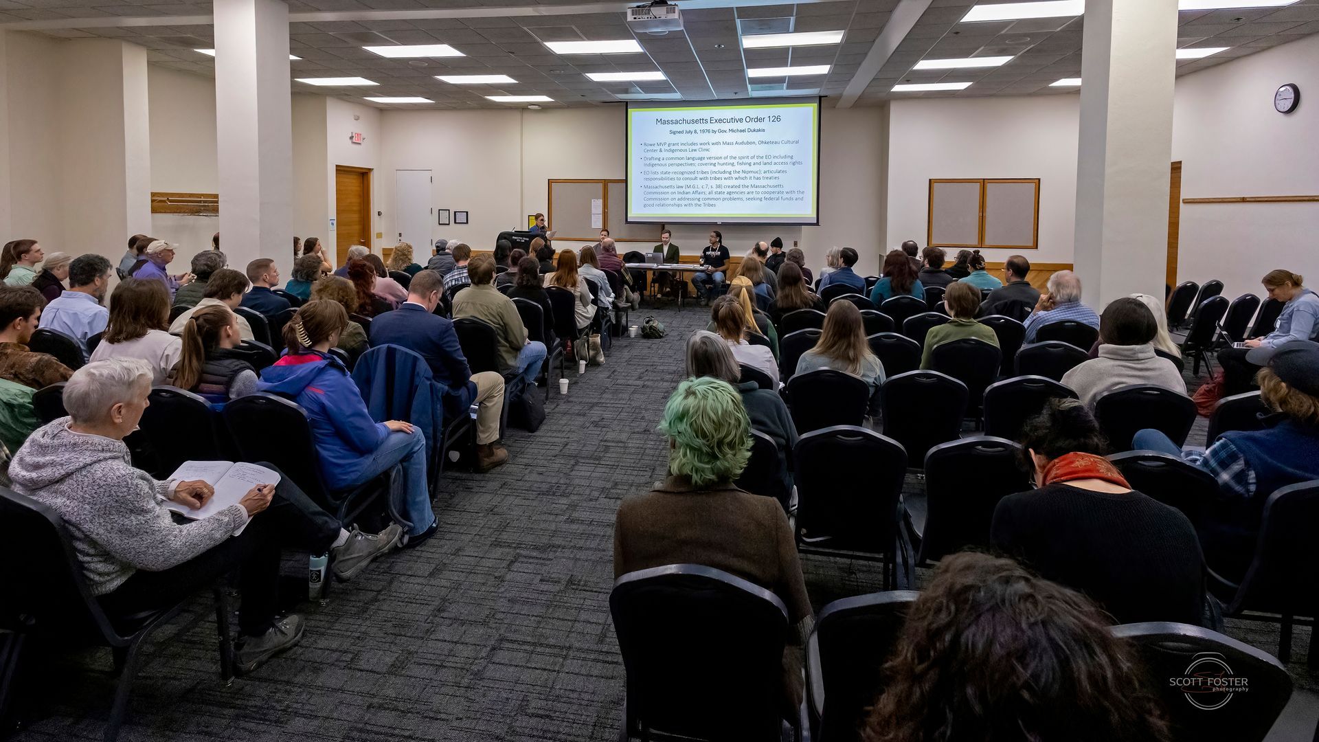 Audience in chairs facing a screen with text. People seated in rows, presentation in a room.