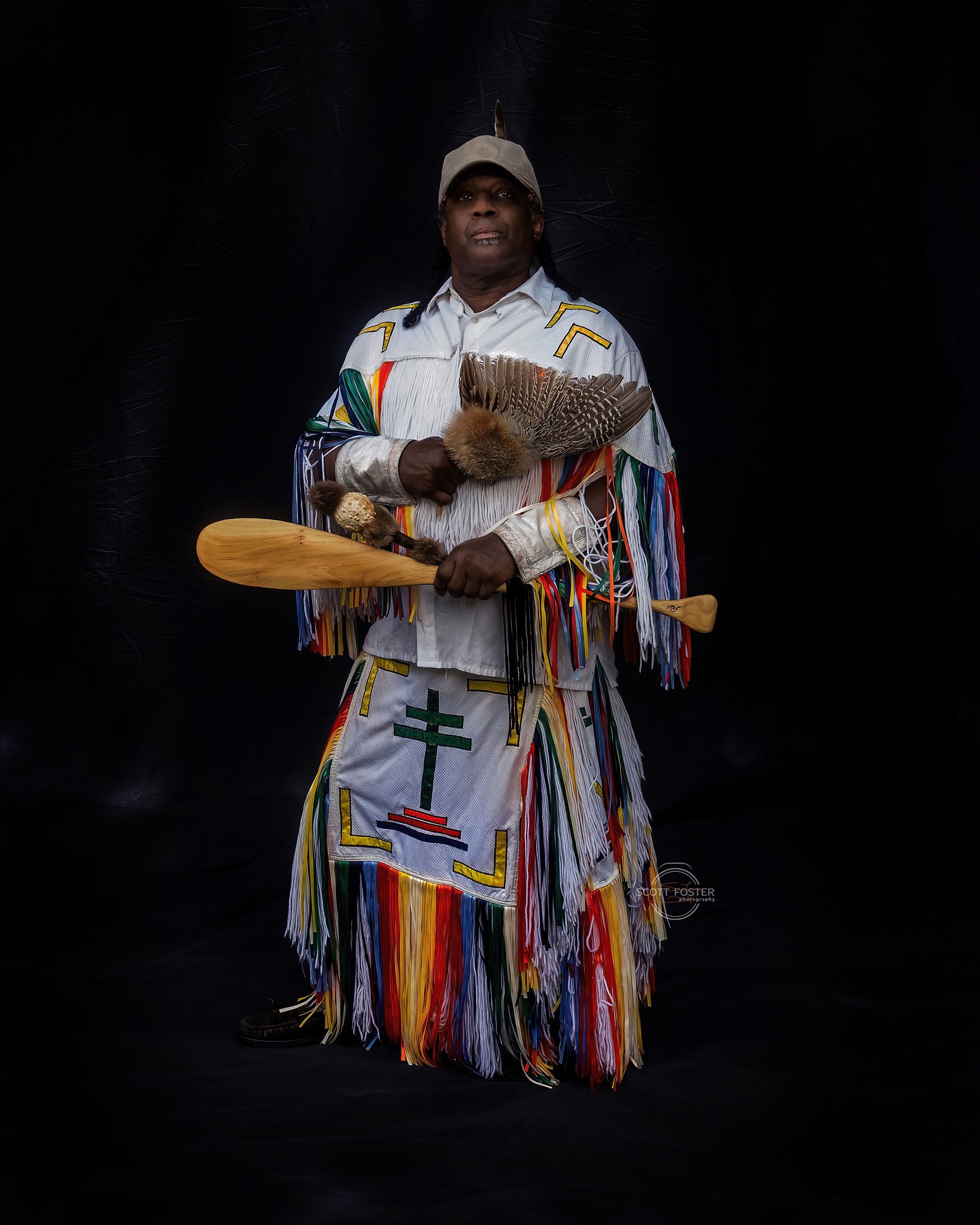 Man in ceremonial attire, holding wooden instruments, standing against a black backdrop.