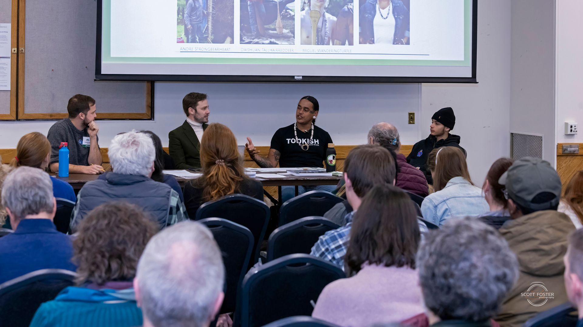 Panel discussion in a room; four people at a table in front of a seated audience, projector screen in the background.