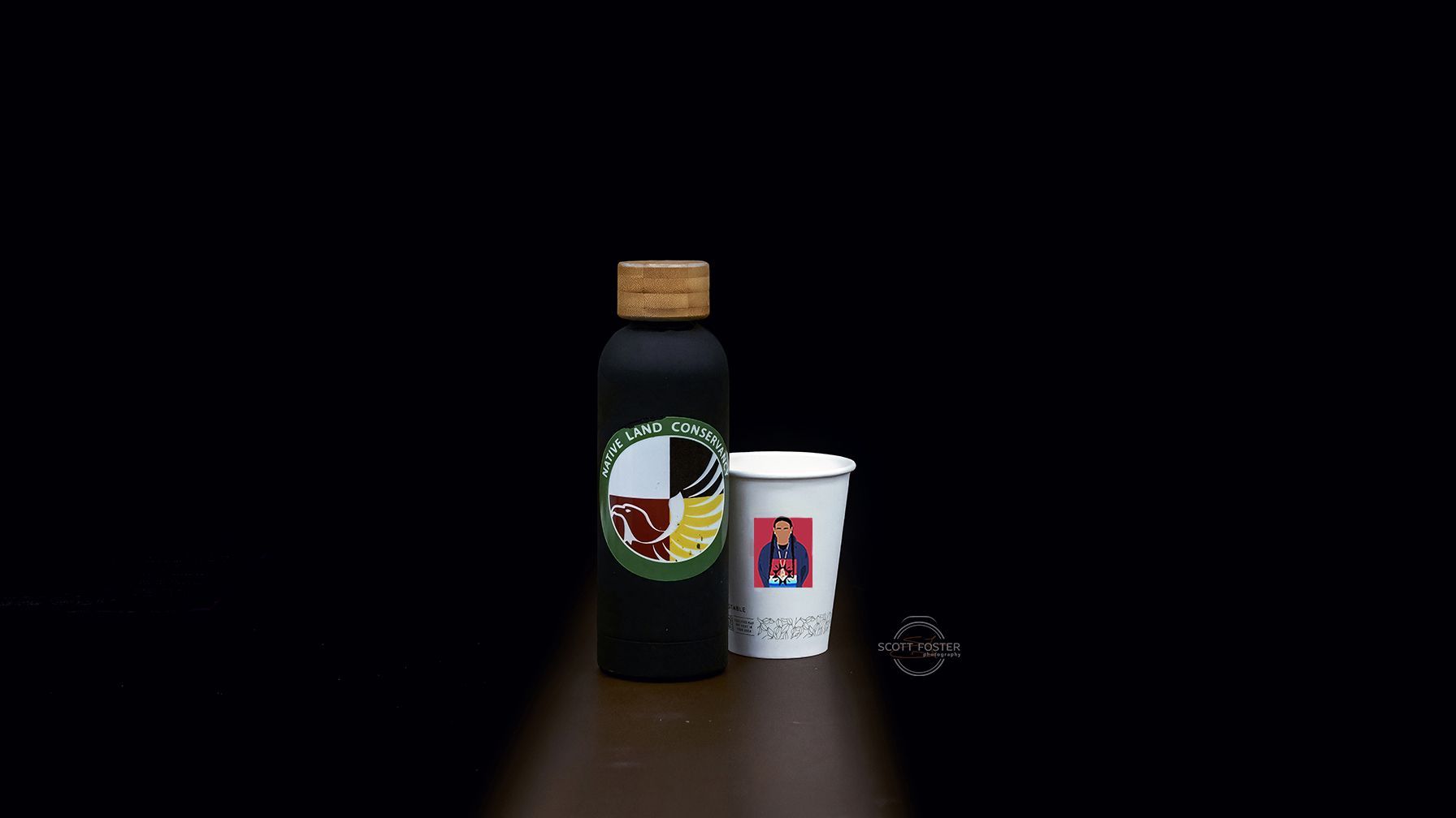 Black water bottle and white cup on brown surface, against a black background.