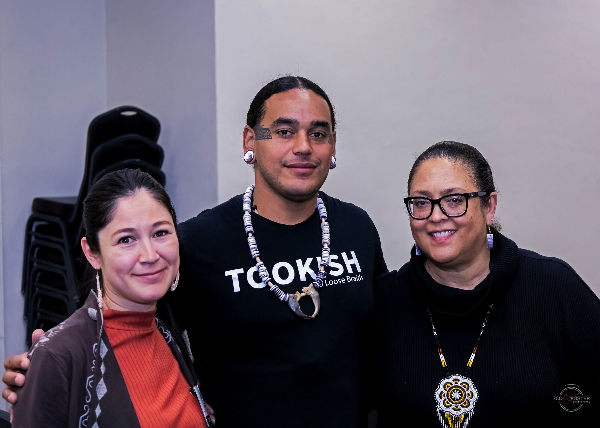 Three people smiling, posing together indoors. Man in black shirt with jewelry; women on either side.