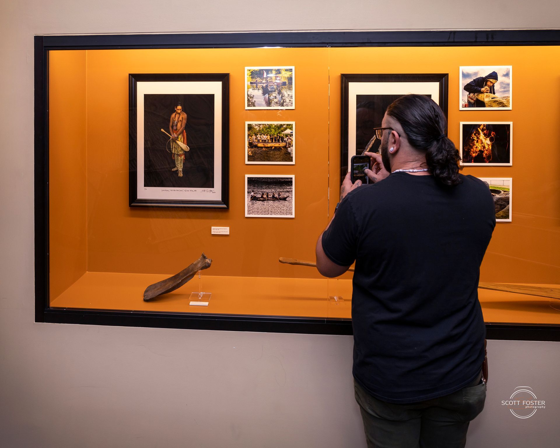 A person photographing artwork in a museum. Display case holds photos and an artifact.