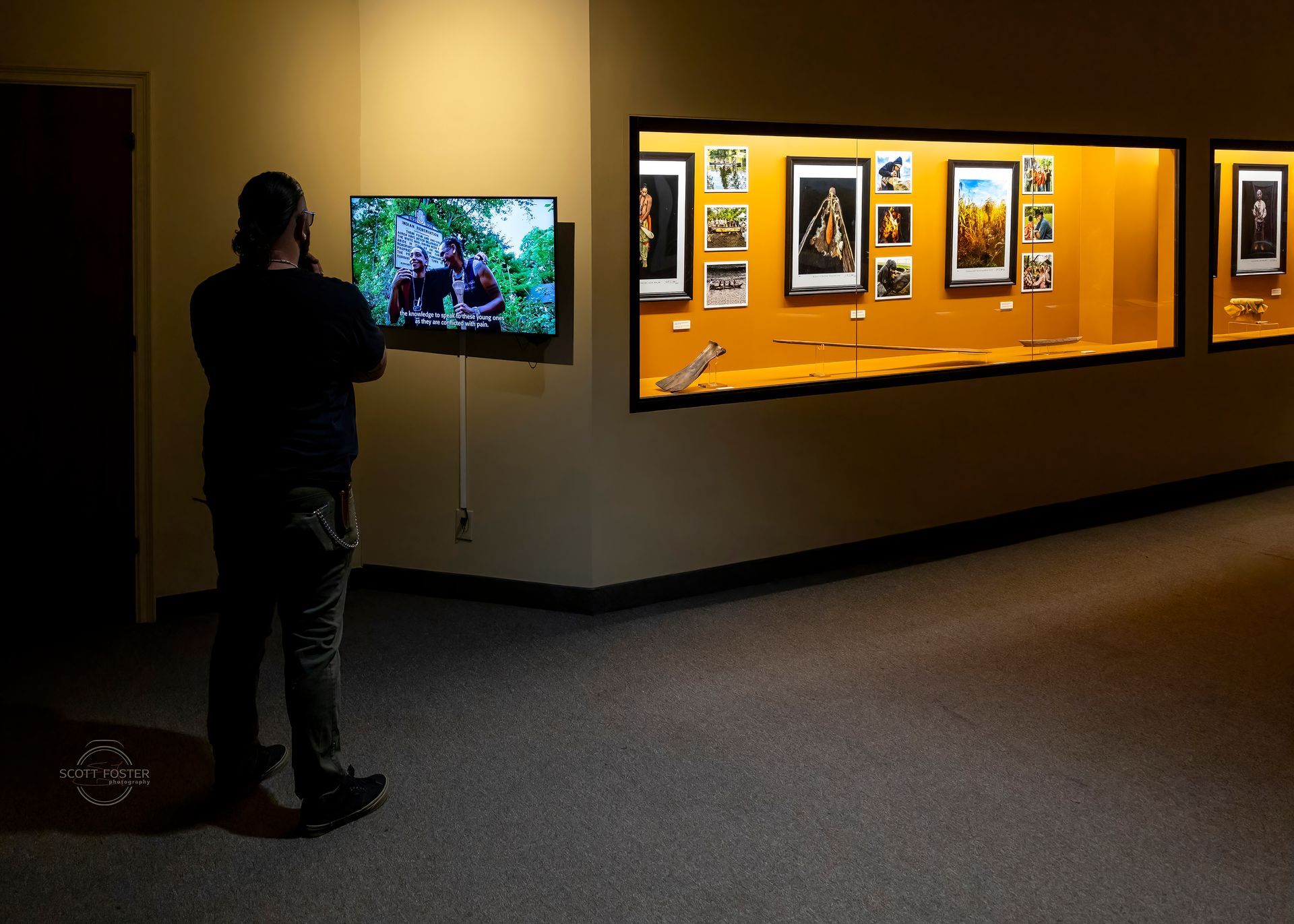 Person in museum looks at a TV screen, next to a display case filled with exhibits.