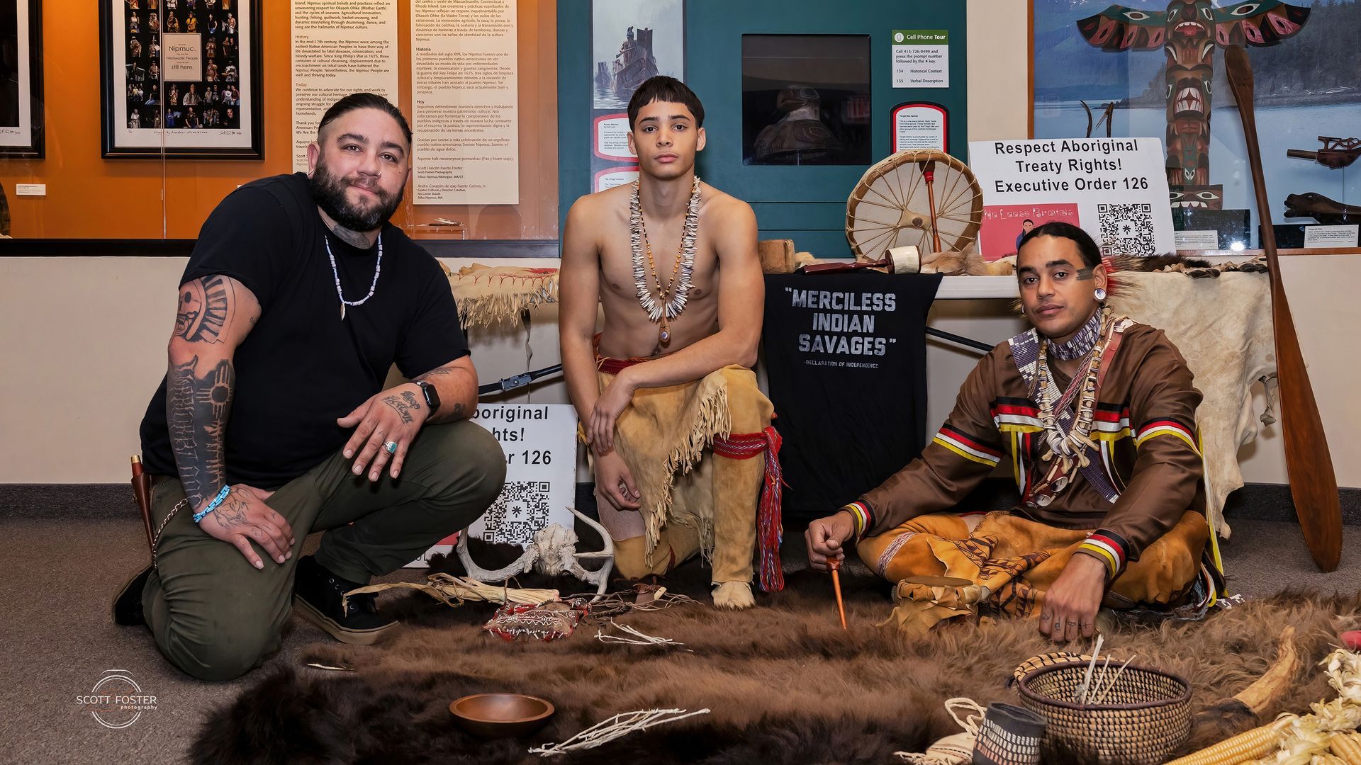Three people in indigenous attire at a display with artifacts. One kneels, two sit.