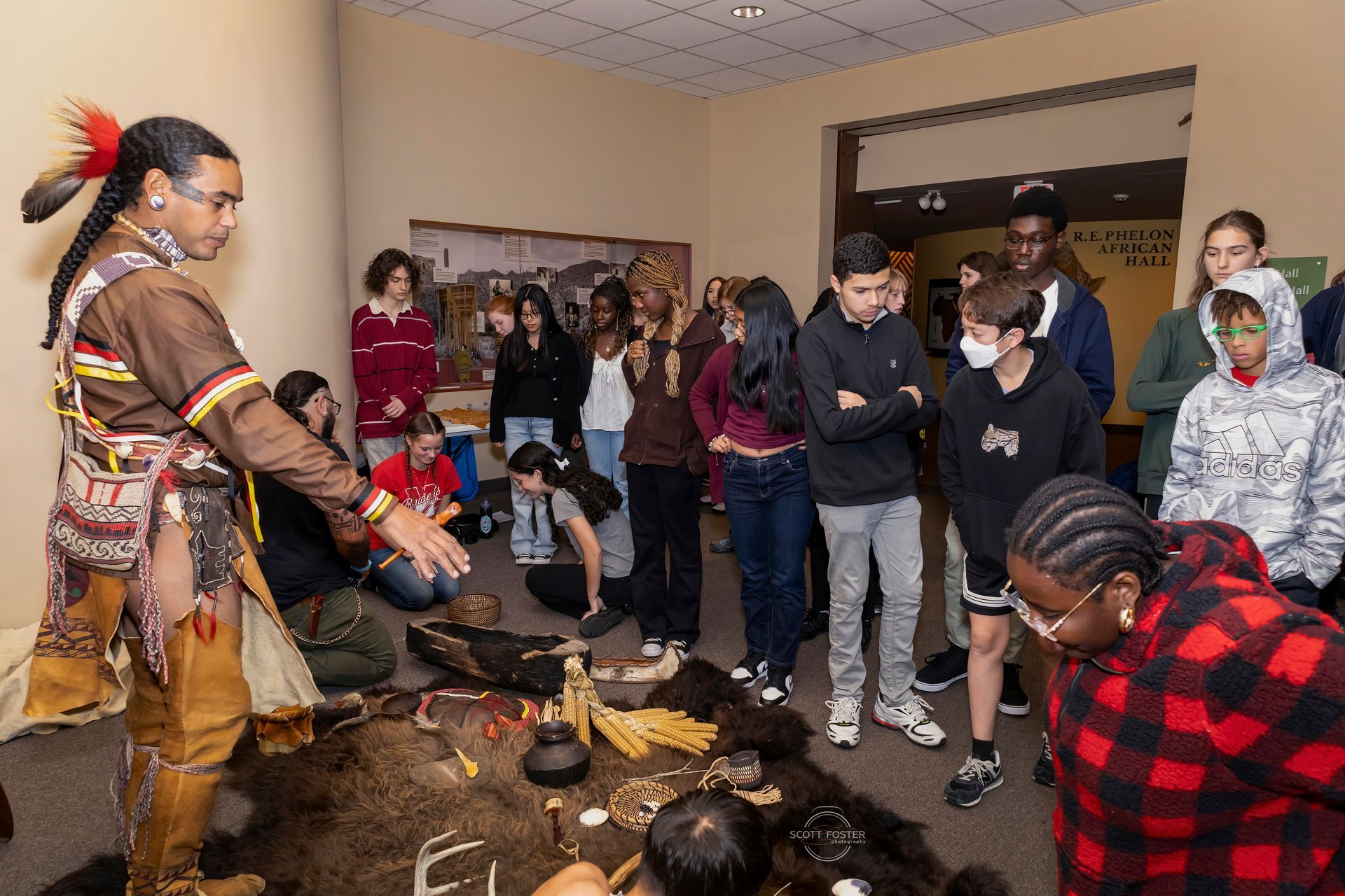 Native American in traditional clothing gestures to a group of students in a museum.