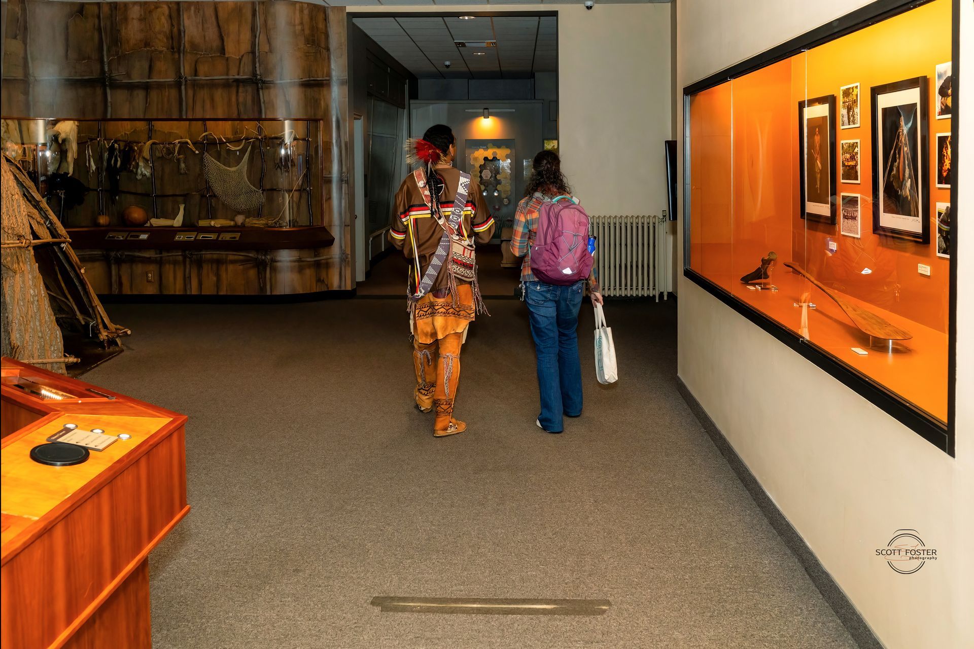 Museum exhibit: Person in Native American attire walks with a visitor; display cases line walls.