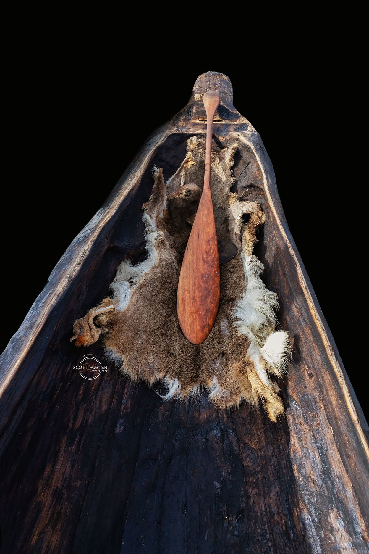 Wooden canoe interior with paddle resting on animal fur. Dark brown and orange hues.