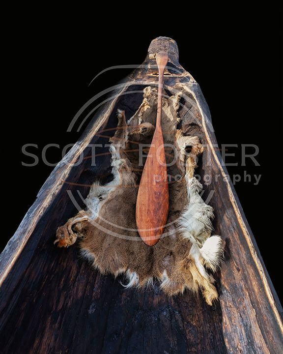 Wooden canoe interior with a wooden paddle atop a fur pelt, black background.