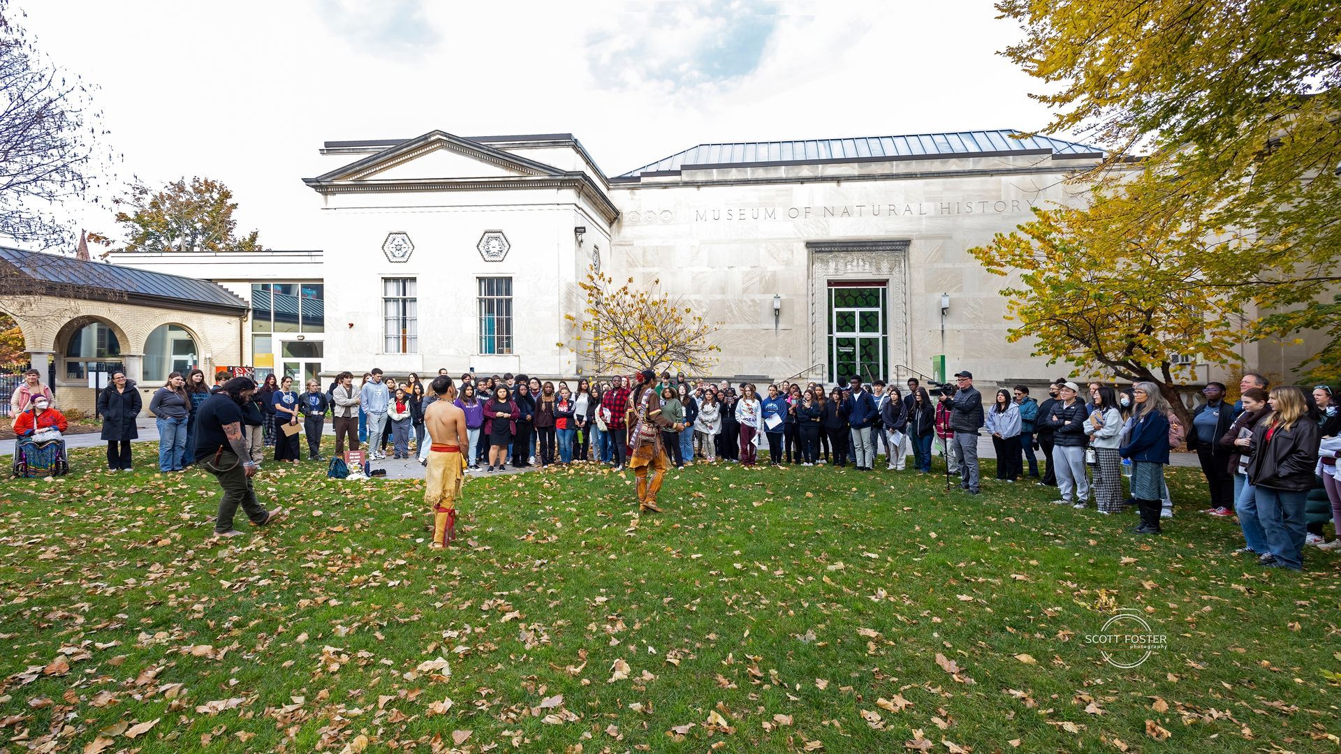 People gathered outside a light-colored building. Some are looking at two people dancing on the grass.