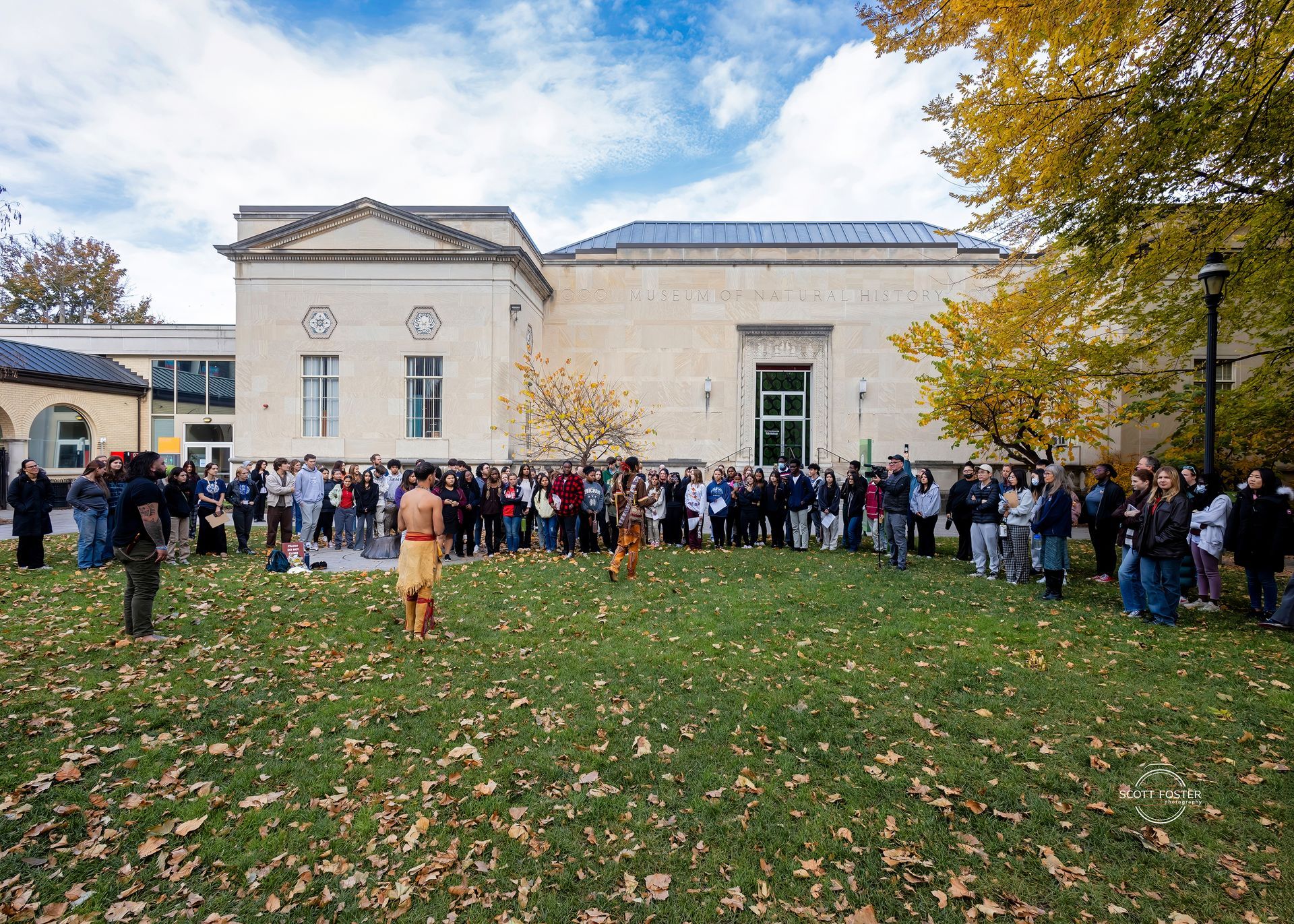 Ceremony with crowd in front of building; two people in traditional garb perform on a lawn.