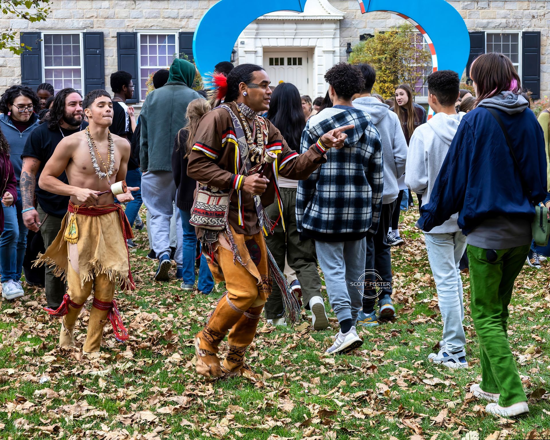 Two Native American dancers perform outdoors, surrounded by a crowd, in front of a building.